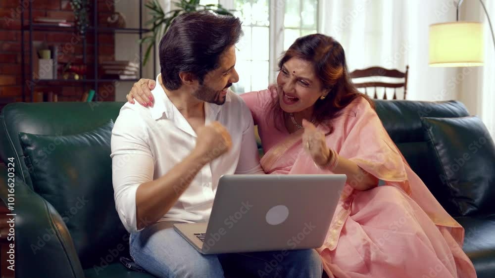 Elderly Indian mother in her sixties and her handsome young son using a laptop together for online shopping or video calling, sitting in a cozy, modern living room with warm, moody lighting