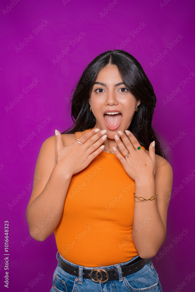Cheerful woman in orange top posing with surprised expression, hands near face, purple background Cheerful woman in orange top posing with surprised expression, hands near face, purple background