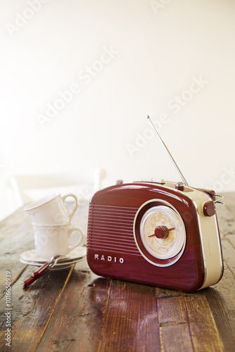 A vintage radio sits on a wooden table beside porcelain white mugs, bathed of morning light. It's time for tea. Vertical photo.