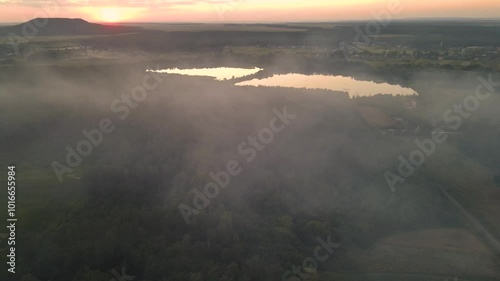 Flying through the fog with an aerial view of a small picturesque town with a diverse landscape: dense green forest, several small lakes or ponds, valleys and a mountain with a bright sunset