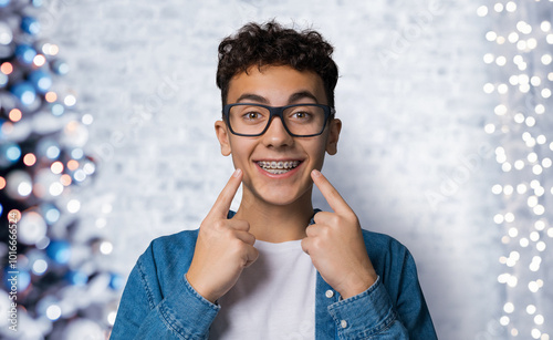 Dental dent care ad concept image - black сurly haired funny young man wear metal braces, glasses, show point white teeth smile. Christmas xmas x-mas tree lights background. Positive optimist concept.