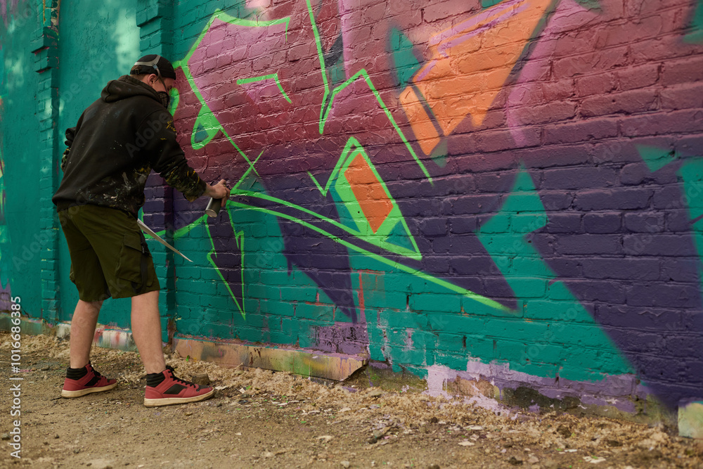 Fototapeta premium Young man in casualwear bending forwards and spraying purple paint on brick wall of building while working over graffiti artwork