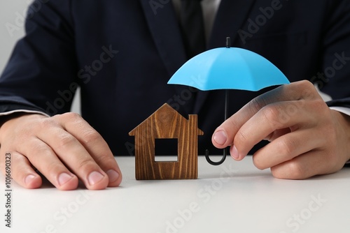 Real estate insurance. Man holding small umbrella above wooden house figure at light table, closeup