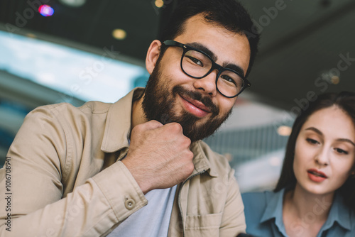 Portrait of cheerful bearded hipster guy in eyewear smiling at camera and enjoying of spending leisure time with beautiful caucasian girlfriend sitting near and looking on screen of cellular phone