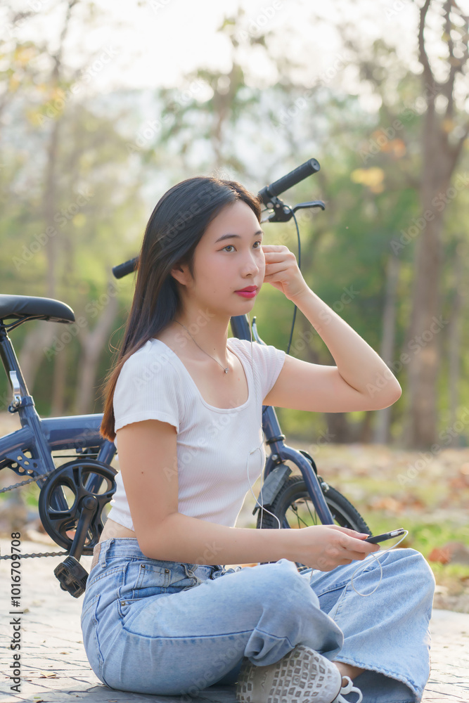 Obraz premium Young asian women wearing earphone and listening audio music on smartphone with happiness while sitting on the street to relaxation with journey travel lifestyle after cycling bicycle in the park