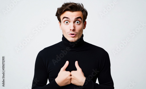 A surprised young man, wearing a black turtleneck, points at his chest with a shocked expression, isolated on a white background. photo