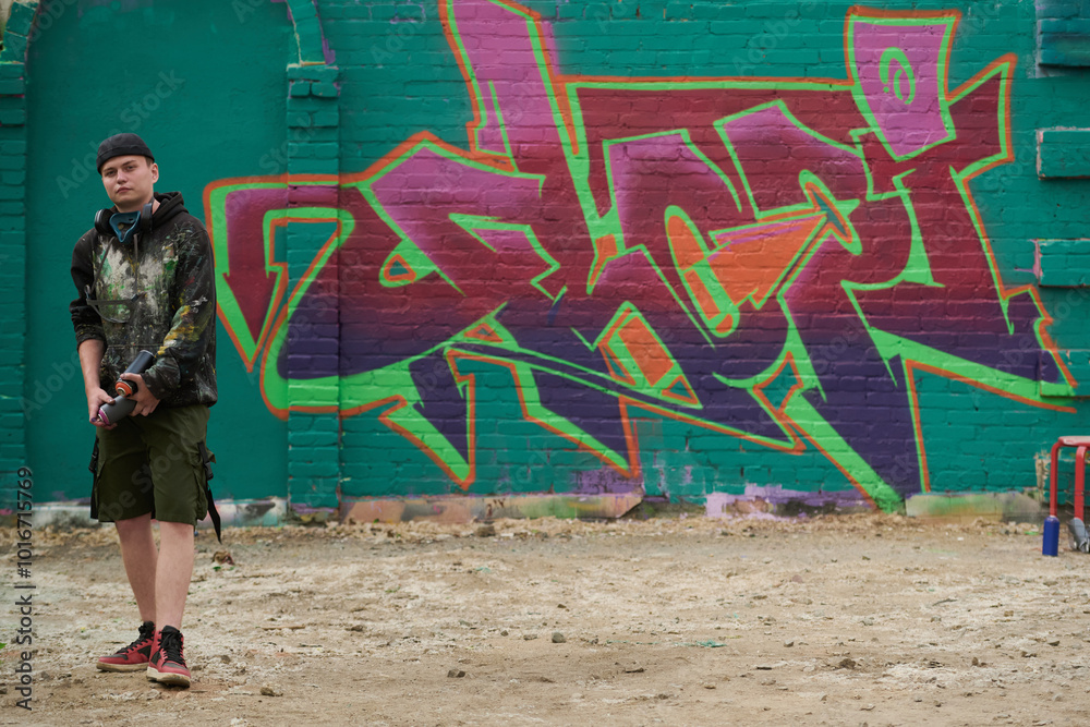 Fototapeta premium Graffiti artwork on green brick wall of building and young man in casualwear holding cans with paints and looking at camera