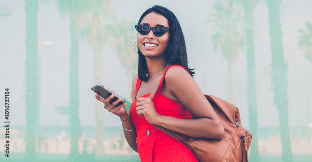 Obraz premium Half length portrait of cheerful woman in fashionable sunglasses holding mobile phone for chatting in hands and laughing at camera during day for walking, gladden jubilate hipster girl phoning