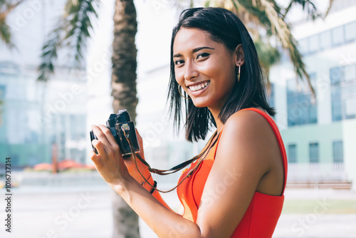 Half length portrait of excited female amateur photographer in casual wear standing in city park and smiling at camera while taking pictures of landmark, tourist feeling good during international trip #1016737314