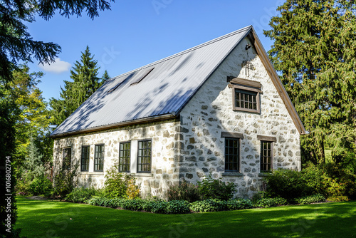 A stone house with an old metal roof stands in the middle of green trees, surrounded by grass and shrubs. The walls are made from white marble or sandstone, creating a rustic charm.