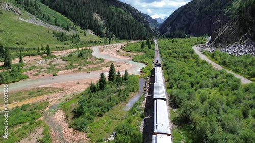 Close up flyover drone shot of scenic passenger train departing Silverton, CO, and approaching a canyon. Flight shows a river in the foreground with mountains in the background.
