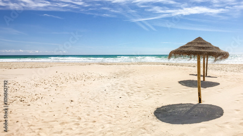 Sunny tropical beach with straw parasols casting shadows on the sand, ideal for summer vacations and relaxation