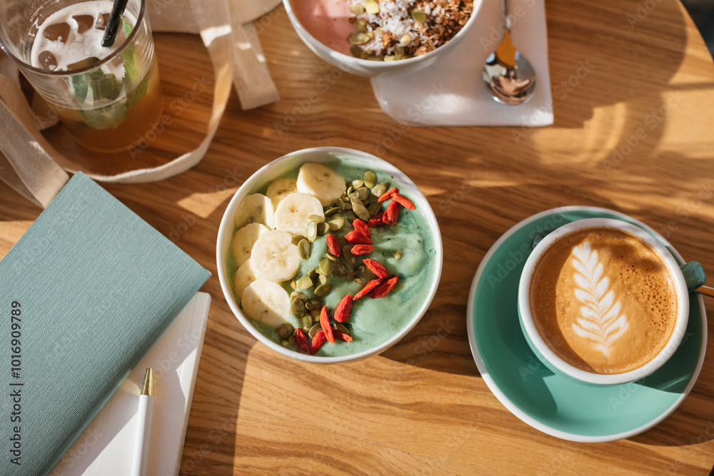 Overhead view of a green smoothie bowl with banana, goji berries, pumpkin seeds, a cappuccino and glass of juice on a table Overhead view of a green smoothie bowl with banana, goji berries, pumpkin seeds, a cappuccino and glass of juice on a table