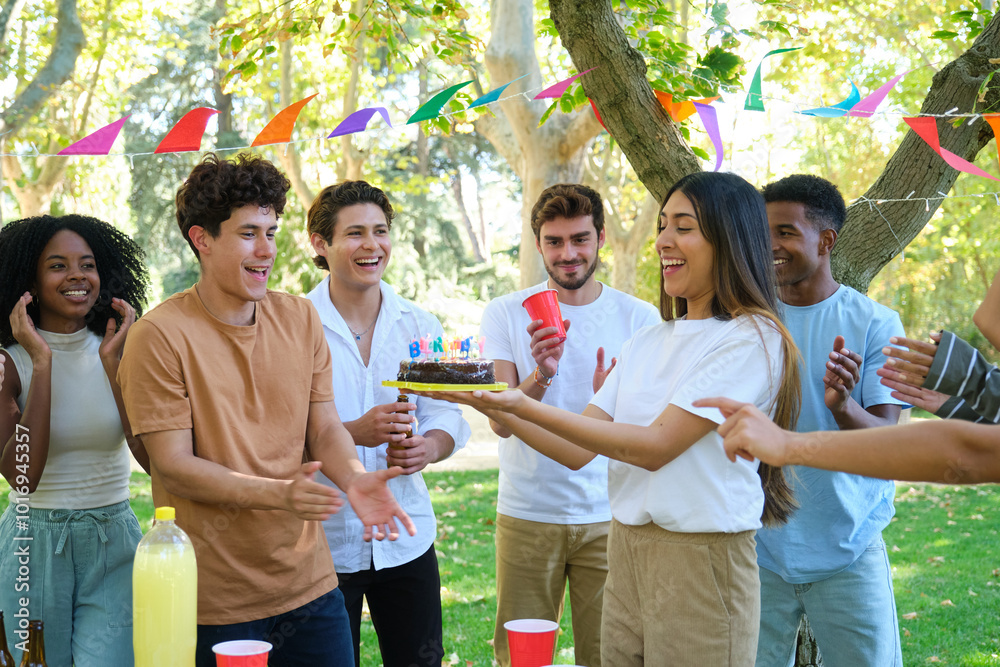 Group of friends celebrating a birthday, a young woman is bringing the cake to the birthday boy, they are in a park