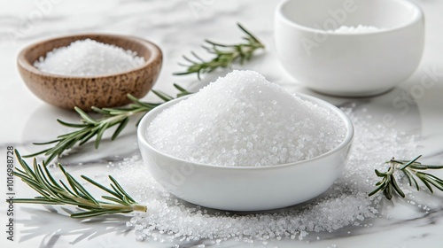 A pile of fine table salt, isolated on a clean marble surface, with decorative sprigs of rosemary and small ceramic bowls