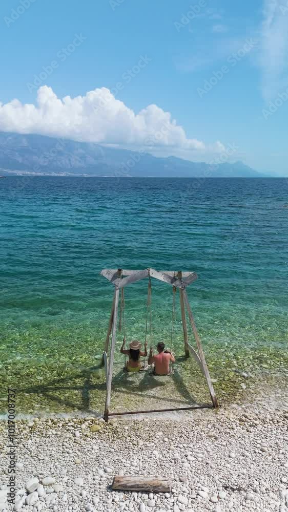 Swaying gently on a rustic swing, two friends relish the tranquil atmosphere of the beach with clear turquoise waters and stunning mountain vistas. Brac Island Croatia Swaying gently on a rustic swing, two friends relish the tranquil atmosphere of the beach with clear turquoise waters and stunning mountain vistas. Brac Island Croatia