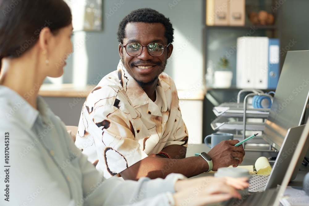 Two people working together in office, focus on smiling African American man talking to colleague while using computer in sunlight Two people working together in office, focus on smiling African American man talking to colleague while using computer in sunlight
