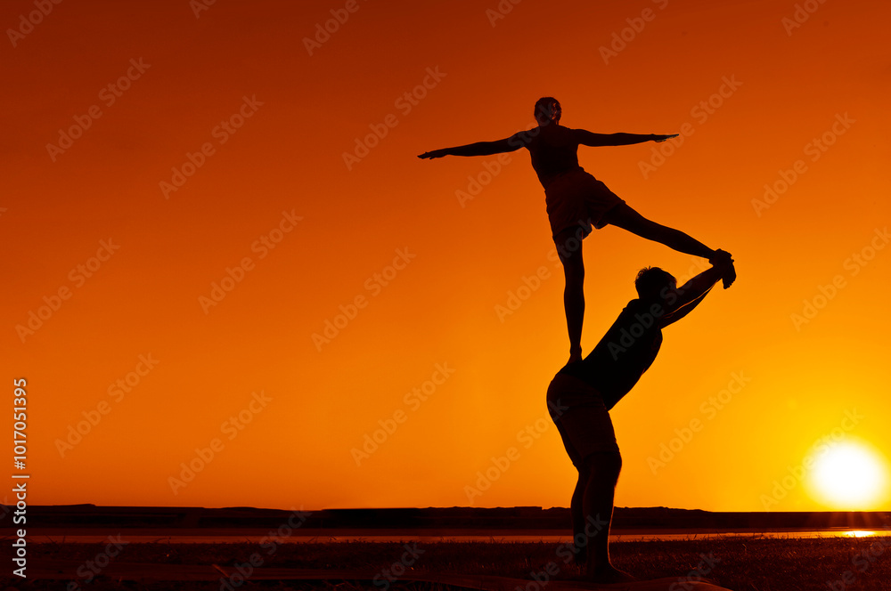 Young adult sports couple doing acro yoga exercises. Silhouette couple practicing yoga at sunset. Slim woman, man do acroyoga. sporty healthy pair doing acrobatic yoga at sunrise. female stand on back Young adult sports couple doing acro yoga exercises. Silhouette couple practicing yoga at sunset. Slim woman, man do acroyoga. sporty healthy pair doing acrobatic yoga at sunrise. female stand on back