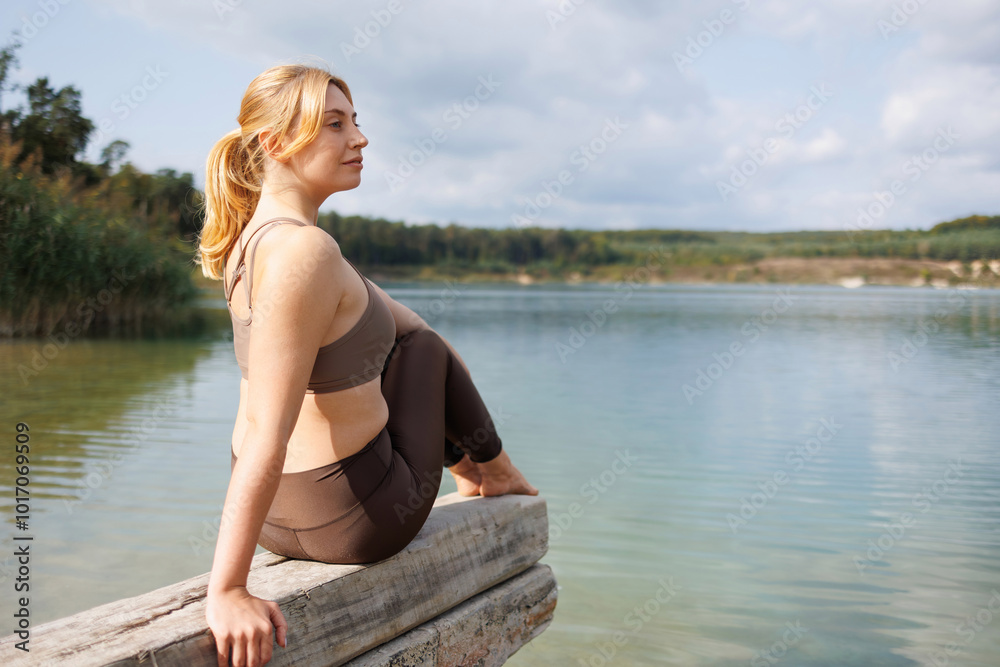 A young woman in sportswear sits on a wooden beam on the shore of a lake A young woman in sportswear sits on a wooden beam on the shore of a lake