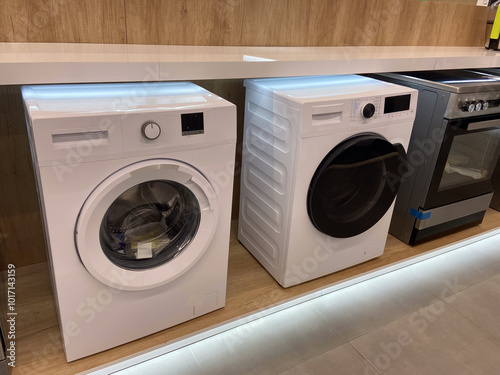 Row of modern washing machines is displayed in an electronics store, showcasing the latest models with sleek designs and advanced features. Under bright lights, the machines stand out against the photo