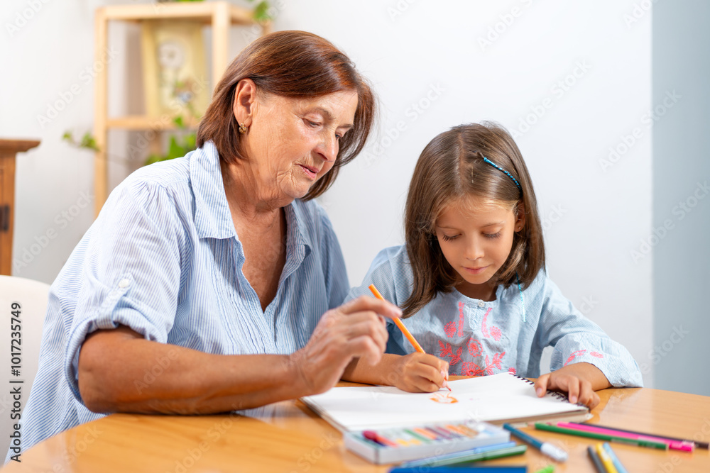 Focused grandmother and granddaughter coloring in book together on table at home. The scene is filled with creativity and warmth, as they bond over their artistic activity Focused grandmother and granddaughter coloring in book together on table at home. The scene is filled with creativity and warmth, as they bond over their artistic activity