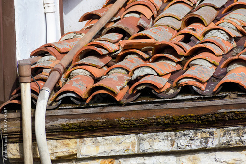 Aged terracotta roof with cracked, misaligned tiles and utility pipes running along the edge photo