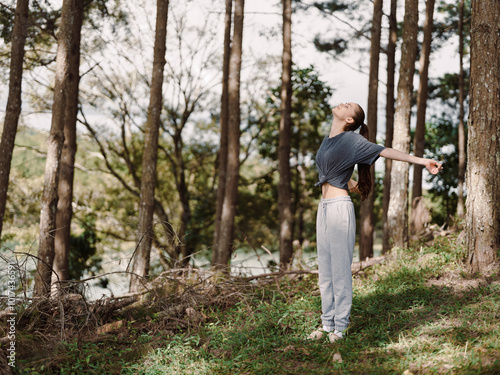 Pleased young woman enjoying fresh air in a forest, arms outstretched, wearing casual sportswear, basking in nature s tranquility Wellness and relaxation concept