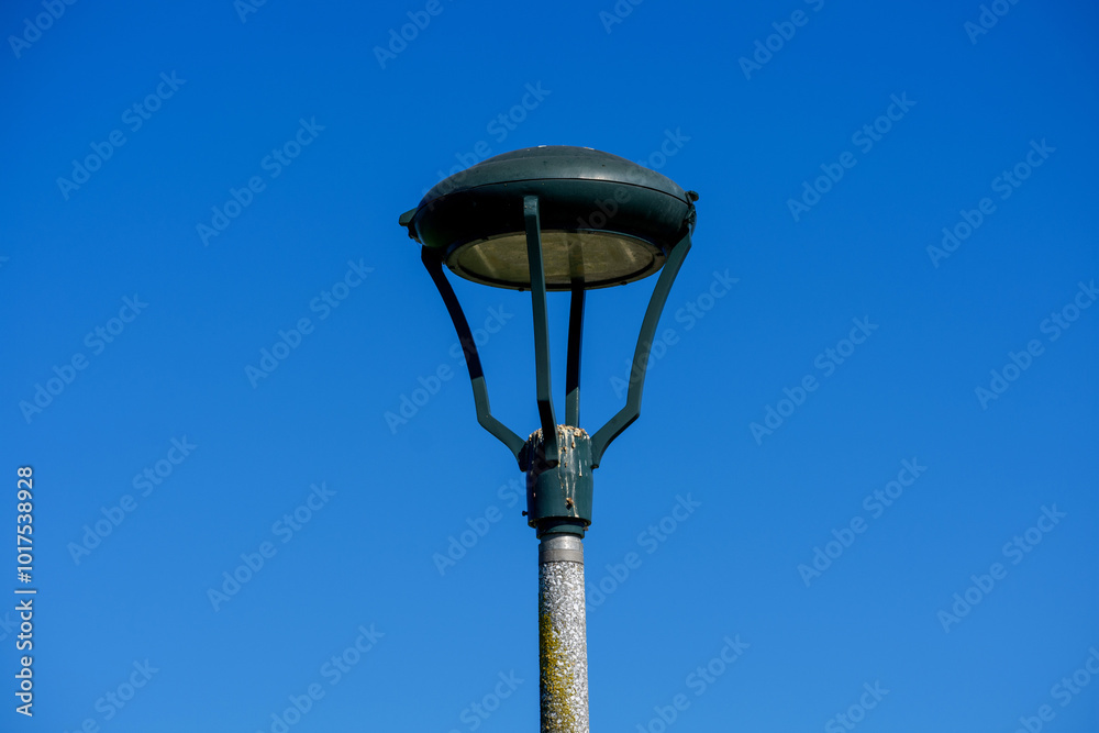 A modern streetlamp with a domed light fixture is set against a bright blue sky. Bird droppings and weathered paint are visible at the base of the lamp head A modern streetlamp with a domed light fixture is set against a bright blue sky. Bird droppings and weathered paint are visible at the base of the lamp head