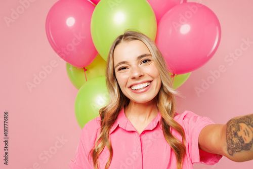 Close up young smiling woman wear shirt celebrating birthday holiday near bunch of colorful air balloons do selfie shot pov on mobile cell phone isolated on plain pink background. Lifestyle concept.