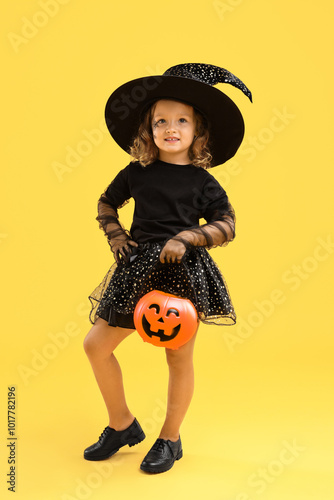 Cute girl with pumpkin bucket dressed like witch on yellow background. Halloween celebration