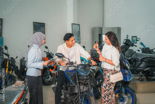 man trying out a new motorcycle and woman giving thumbs up with saleswoman in showroom