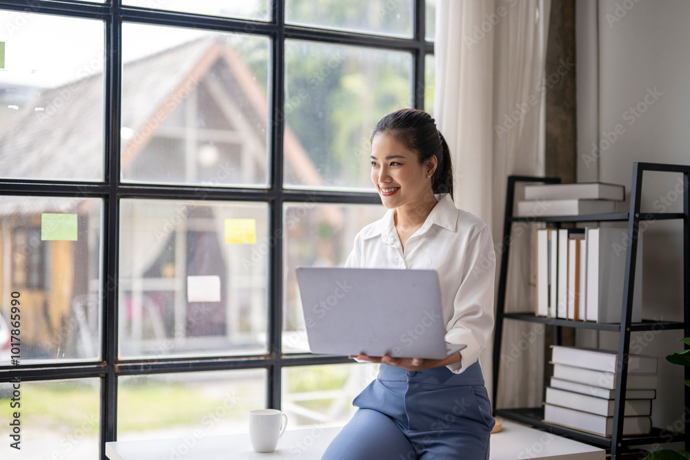 Businesswoman smiling and holding laptop while sitting on desk in home office Businesswoman smiling and holding laptop while sitting on desk in home office