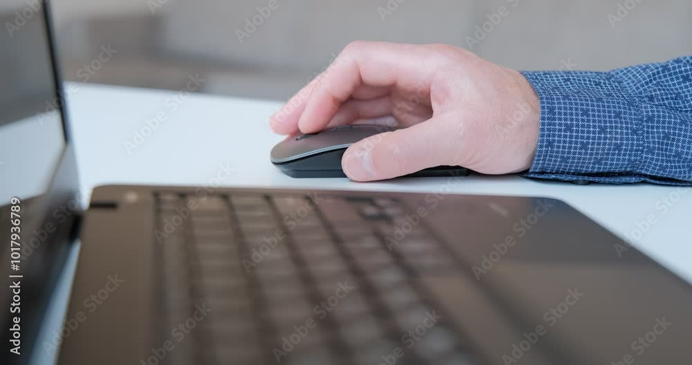 An individual is engaged in work, using a computer mouse to navigate a laptop in a contemporary workspace. The setting is bright and organized, reflecting productivity.