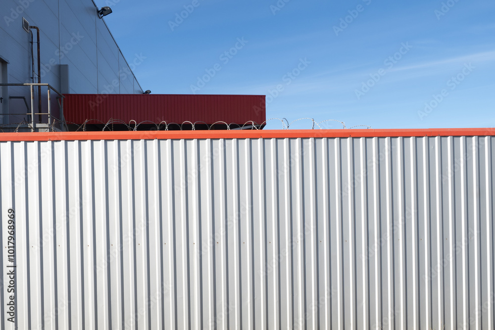 Solid steel fence. Barbed wire over fencing. Fence around industrial building. Territory is fenced with corrugated sheets and barbed wire. Fence made of metal sheet under blue sky. Protection, safety Solid steel fence. Barbed wire over fencing. Fence around industrial building. Territory is fenced with corrugated sheets and barbed wire. Fence made of metal sheet under blue sky. Protection, safety