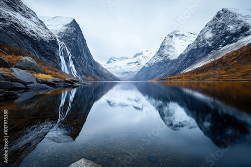 A serene mountain landscape showing a calm lake surrounded by snow-capped mountains, with a waterfall flowing from one of the mountains into the lake.