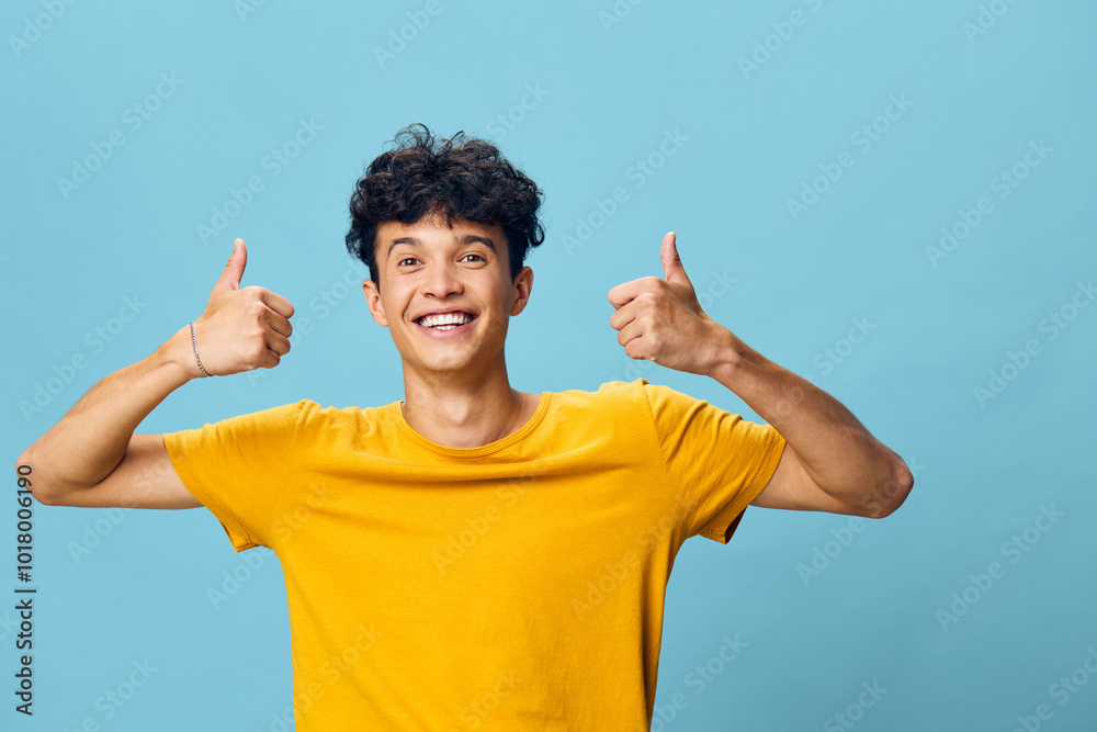 Happy young man showing thumbs up gesture with a bright smile against a light blue background, conveying positivity and enthusiasm, wearing a yellow t shirt Happy young man showing thumbs up gesture with a bright smile against a light blue background, conveying positivity and enthusiasm, wearing a yellow t shirt