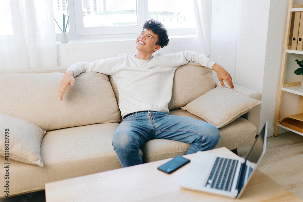 Comfortable young man relaxing on a sofa in a bright, modern living room with a laptop nearby Cheerful expression, casual clothing, showcasing a cozy atmosphere Comfortable young man relaxing on a sofa in a bright, modern living room with a laptop nearby Cheerful expression, casual clothing, showcasing a cozy atmosphere