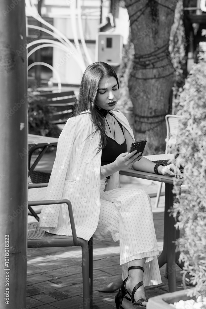 Obraz premium Stylish woman in a monochrome outfit seated at a cafe, engaged with her smartphone under natural light