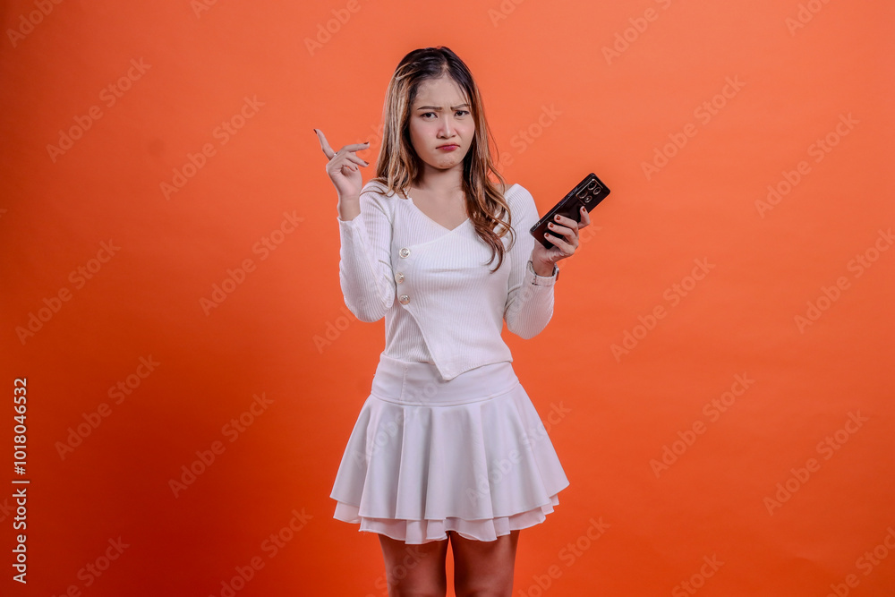 portrait of beautiful adult woman in white top standing pouting to camera holding mobile phone, cellphone and hand pointing on orange background isolation. for technology, promotion, lifestyle portrait of beautiful adult woman in white top standing pouting to camera holding mobile phone, cellphone and hand pointing on orange background isolation. for technology, promotion, lifestyle