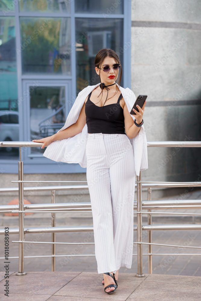 Fototapeta premium Stylish Young Woman in Chic Outfit with Sunglasses and Mobile Phone Outdoors