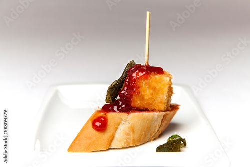 White plate of crispy bread skewer with a portion of breaded cheese and strawberry jam on a white background