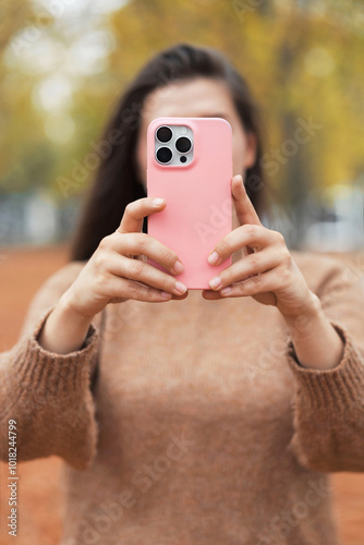 Vertical shot young woman holding smartphone in pink silicone case outdoors. Teen girl take a picture by mobile phone in autumn city park. 