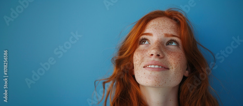 a red haired girl with freckles looking up diagonally, smile expression, blue background.
Redhead, young, woman, female, portrait, smiling, looking up photo