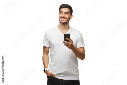 Young man standing in white t-shirt holding smartphone in hand and looking away with dreamful smile