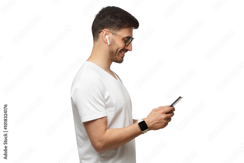Side shot of young man in white t-shirt, wearing smartwatch and wireless earphones, holding phone and smiling at screen Side shot of young man in white t-shirt, wearing smartwatch and wireless earphones, holding phone and smiling at screen