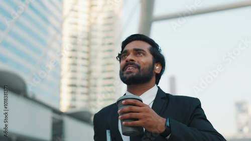 Inspired hindu male walking by downtown district and looking around at skyscrapers in morning. Corporate worker listening to music through earphones on way to office and drinking coffee on go.