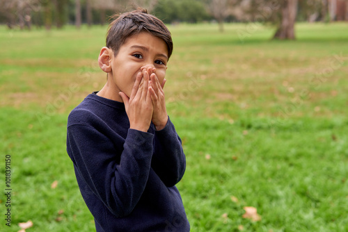 latino boy with a mischievous face covering his mouth and looking at the camera after saying a rude word, in the background a green forest.