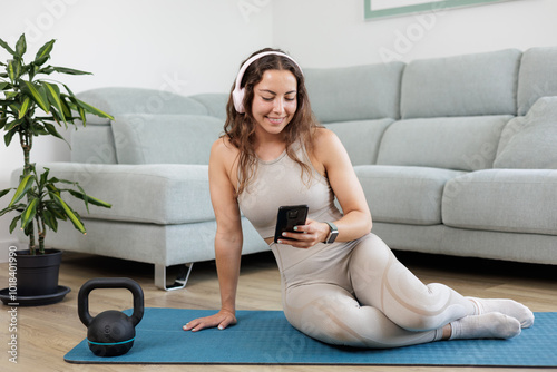 Young woman searches on her mobil phone for exercises to do from home with a kettlebell and listens to music with pink headphones. Online personal trainer.