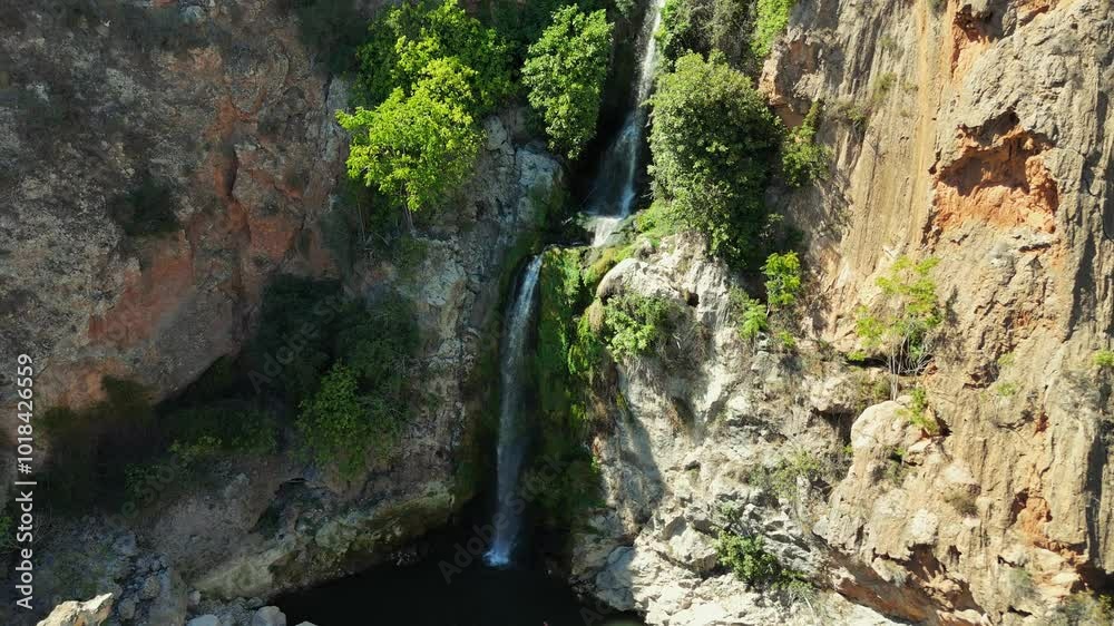 Drone Shot, Camera panning across beautiful waterfall flowing over steep cliff into small pond. Lush green trees and vegetation surround the waterfall. Aerial view Drone Shot, Camera panning across beautiful waterfall flowing over steep cliff into small pond. Lush green trees and vegetation surround the waterfall. Aerial view