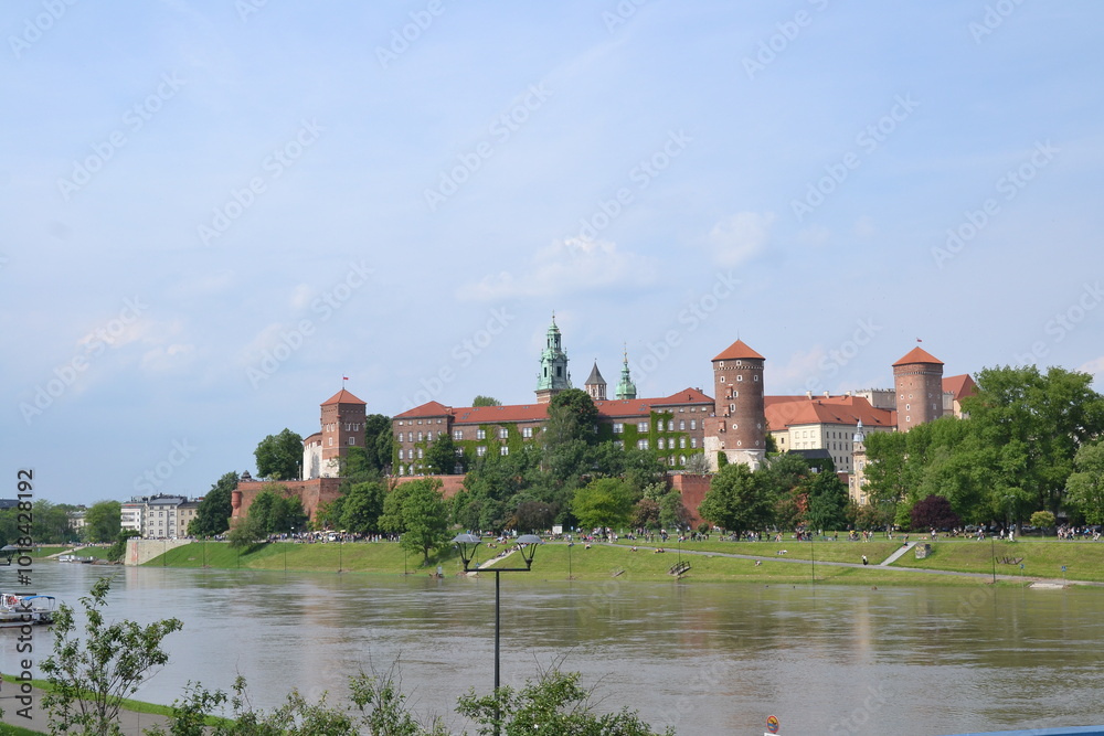 Fototapeta premium A scenic view of Wawel Castle in Kraków, Poland, showcasing its historic red-brick architecture, green domes, and surrounding park along the Vistula River under a clear sky.